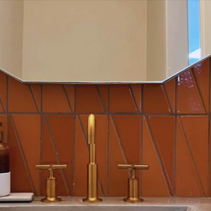 A bathroom sink with gold fixtures is set against a wall of orange geometric tiles. A white pot with orchids and a soap dispenser sit on the countertop. A large mirror hangs above the sink, reflecting light.